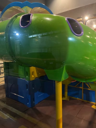 The image depicts a green, tube-shaped children's slide with purple accents, likely located on a playground. The slide is connected to a blue and yellow play structure. The background includes a tiled floor and elements of the building interior. The location is Bencongan Indah, Indonesia. No people are visible.