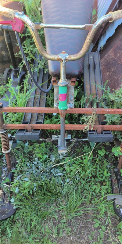 The image shows a close-up, slightly elevated view of a weathered, multi-colored object, possibly a piece of gardening equipment or an old bicycle frame, situated outdoors in a grassy, overgrown area. The object has a rusty metal frame, a black seat with red stitching, and a green and pink painted vertical support. The foreground is dominated by green grass and weeds, with some dried foliage. In the background, there are glimpses of other metallic structures and possibly more vegetation. The scene appears to be in a rural or unkempt urban setting. The specific location in the city of Есипово, Russia cannot be determined from the visual information provided in the image. There are no people or animals visible in the frame.