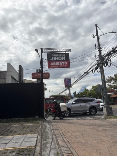 The scene is a street corner in San Rafael, Costa Rica (latitude: 9.938, longitude: -84.153), under an overcast sky. A man in a black helmet and red jacket is maneuvering a red motorcycle with a white box attached to the back out of a parking space. The motorcycle is turning left onto the street. He is in front of a black gate, partially obscured by a silver SUV that is in the process of pulling out onto the street. Another vehicle, a red van with a roof rack, is parked on the street. A sign on a pole reads "COLCHONES JIRON ABIERTO De Lunes a Domingo," indicating a mattress store. There is a utility pole with numerous wires crossing and connecting to it. The sidewalk is paved with small, brick-like blocks, and there are yellow lines painted on the ground. The street is paved with cobblestones.