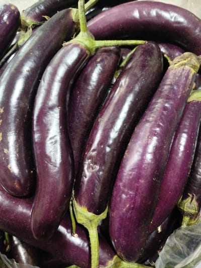 A close-up, overhead shot shows a pile of dark purple eggplants. They are long and slender, with smooth, glossy skin. Several eggplants have green stems attached. The eggplants are densely packed, suggesting they are being displayed or sold at a market. The lighting is bright, highlighting the sheen of the eggplants and casting subtle shadows. The overall impression is one of fresh produce. This image was likely taken at a local market in Keudelapang, Indonesia, a common location for such produce displays.