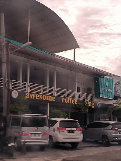 This image captures a street scene in Jurong Peukan, Indonesia, depicting a row of commercial buildings with parked vehicles in the foreground. The sky is overcast with pale, pinkish clouds, suggesting either early morning or late afternoon, under daylight conditions.

The prominent structure is a multi-story building featuring a distinctive large, curved metal roof over the top floor. Below this, a long, white-railed balcony spans across the second floor, supported by pillars. Ceiling fans are visible on the second-story balcony.

On the ground floor, large golden letters spell out "awesome coffee", indicating a café or coffee shop. Further along the building to the right, a prominent teal-colored sign with a tooth logo displays "RZ DENTAL", signifying a dental clinic. A partial sign that reads "fadra" is also visible to the far right.

Several cars are parked along the street in front of the buildings. In the left foreground, the rear of a silver van or MPV is visible, with a license plate reading "B 2330 PX". To its right, a white SUV is parked, and further right, another silver SUV is seen. A street light with a horizontal lamp extends from a pole on the left, and power lines crisscross the upper part of the image. Small trees and bushes provide some greenery along the storefronts. No people are clearly identifiable in the scene, which appears to be a typical urban commercial area.