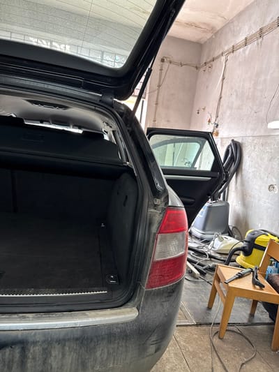 This image depicts a black station wagon, likely a Skoda Octavia Combi, with its tailgate and rear passenger side door open, situated inside a utilitarian building, likely a garage or vehicle detailing shop, in Gornik, Bulgaria. The vehicle's cargo area is empty, revealing a dark carpeted floor and a partially retracted cargo cover. The rear bumper displays dirt and superficial scuff marks on its passenger side. The car's interior, visible through the open rear door, appears dark.

The surrounding environment consists of light-colored, rough concrete or plaster walls with exposed electrical conduits running horizontally and vertically. The floor is concrete, covered with a network of electrical cables and hoses. To the right of the vehicle, a yellow Karcher wet/dry vacuum cleaner is visible on the floor, alongside a larger, gray industrial cleaning unit with coiled black hoses. A light wooden chair is positioned between the car and the wall, with two dark-colored cleaning attachments resting on its seat. An overhead fluorescent light fixture is partially visible in the upper right. No individuals are present in the scene.