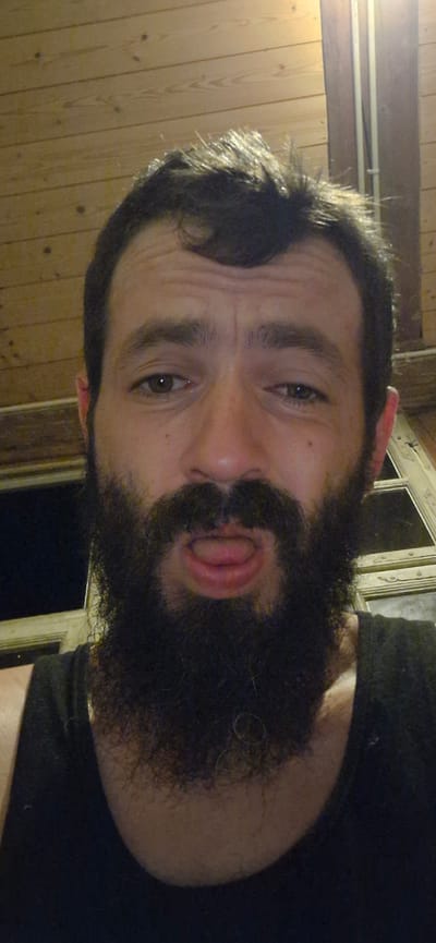 The image shows a close-up, selfie-style photograph of a man with dark hair and a full beard, taken indoors. The man's face is centered in the frame. He has a light complexion, dark eyebrows, and brown eyes. His tongue is slightly protruding from his mouth, and his lips are slightly parted. He is wearing a dark tank top. The background consists of wooden paneling on the ceiling and a wall, with a partial view of a window or door frame with horizontal panes and vertical elements. There are some light fixtures visible in the upper right portion of the image. The overall lighting suggests an indoor setting, possibly at dusk or night given the darkness outside the window. There is no discernible indication of the man's location within Wattenwil, Switzerland.