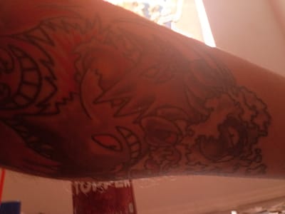 A close-up, indoor photograph displays a tattoo on an arm, presumably human. The tattoo features cartoon-like depictions including what appears to be a grinning character with large teeth and red eyes, possibly a monster or a caricature. Another figure appears to be on the left of the frame. The location is provided as Clarksville, United States of America.