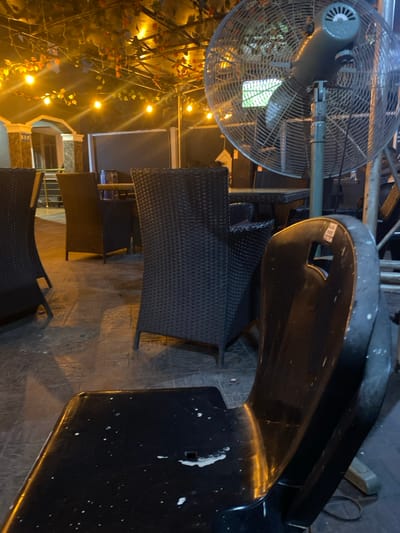 This image depicts an unoccupied outdoor dining or lounge area within Igbessa, Nigeria, observed during nighttime hours. The immediate foreground features the backrest and partial seat of a black plastic chair, marked with numerous white splatters. In the midground, multiple dark-colored, wicker-style chairs are arranged around glass-topped tables. A large, grey industrial standing fan with a circular metallic cage is positioned prominently on the right side of the frame. Overhead, an open trellis or canopy structure is draped with artificial foliage, from which multiple spherical string lights emit a warm, orange glow. The ground beneath the seating area is paved with dark-colored, possibly tiled, material. In the background, dimly lit architectural elements, including decorative pillars and windows, indicate a building structure. No individuals are present within the captured scene.