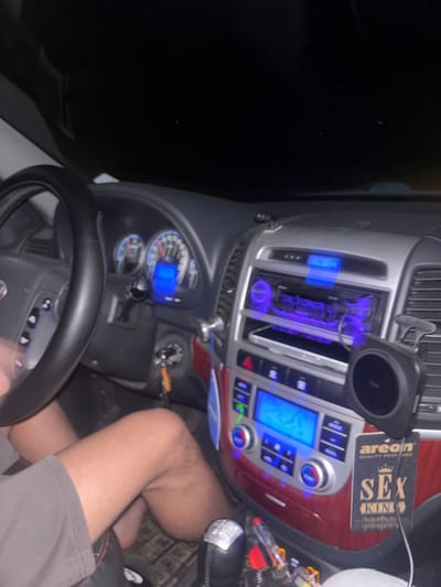 The image depicts the interior of a car at night. The dashboard is visible, featuring blue illuminated displays for the radio and climate control. An air freshener labeled "SEX KING" hangs nearby. The steering wheel is in the foreground. A person's leg, clad in shorts, is also visible. The location, Gromshin, Bulgaria, cannot be verified from the image content.