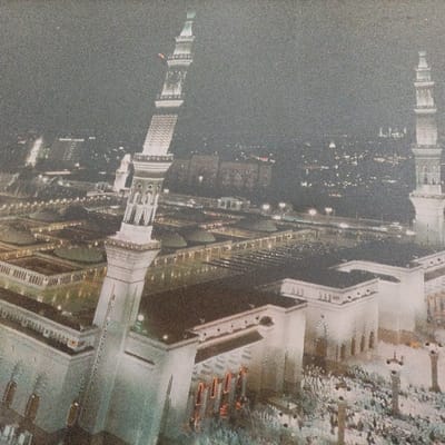 This grainy, high-angle nocturnal image depicts a vast, brightly illuminated mosque complex, identified as being in the city of Pasir Muncang, Indonesia. The central structure features a large, flat roof adorned with numerous domes, surrounded by expansive courtyards. Several ornate, multi-tiered minarets, vertically prominent and incandescently lit, punctuate the skyline; one minaret dominates the left foreground, while another is visible on the right. A massive congregation of people, appearing as dense clusters of light-colored figures, occupies the open areas and courtyards surrounding the mosque, indicating a large-scale religious gathering. In the distant background, the ambient illumination from a sprawling urban landscape extends under a dark sky, with scattered city lights marking buildings and thoroughfares. The overall scene is static, capturing the architectural grandeur of the mosque and the presence of a multitude of worshippers under artificial light.