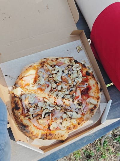 A pizza rests in an open cardboard box on a dark wooden surface, likely a table. The pizza is round and appears to be thin-crust, with toppings of sliced mushrooms, ham, and cheese, along with a red sauce visible underneath. Some areas of the crust are charred, indicating it may have been cooked in a wood-fired oven. The box is lined with white paper. To the right of the box, a person wearing a bright red garment, likely pants or a skirt, is partially visible. To the left, a sliver of blue denim, possibly jeans, is also in view, suggesting someone is seated or standing near the pizza. The scene is outdoors, with some green vegetation and dry ground visible in the background. Sunlight casts shadows, suggesting it is daytime, possibly late morning or afternoon. The overall mood seems casual and relaxed, hinting at a meal being enjoyed outdoors. There is no visible text in the image.