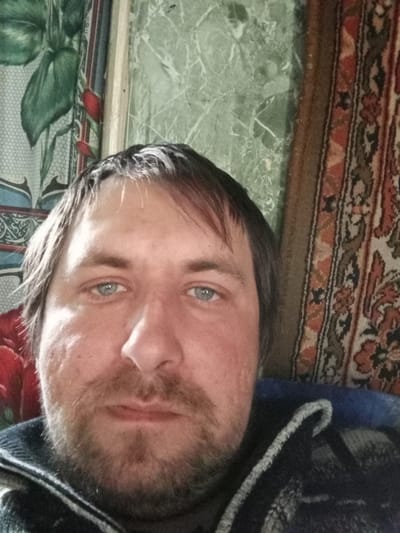 The image depicts a man with light blue eyes, dark hair, and stubble. He is wearing a dark, possibly patterned, sweater or jacket. Behind him are three distinct wall coverings or hangings. The leftmost appears to be a floral pattern, with large green leaves and red flowers. The center is a light green marble-like pattern, and the rightmost is a tapestry with an intricate, multi-colored design. There is no definitive information in the image to pinpoint a specific location in Parabel, Russia, or any city.