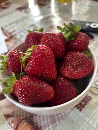 A close-up, high-angle shot features a white bowl overflowing with ripe strawberries. The strawberries are a deep red, with visible seeds and green stems and leaves. The bowl is placed on a tablecloth with a checkered pattern and a floral design. In the background, a remote control and part of a bottle are visible, suggesting an indoor setting. The scene is brightly lit, with some glare reflecting off the strawberries and the tablecloth. The word "SUCRE" (French for sugar) is partially visible on the rim of the bowl. This image was taken in Zubov, Russia.