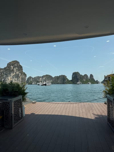 The image depicts a scenic view of Ha Long Bay, located in Tuan Chau Ward, Vietnam, from a slightly elevated perspective. A body of water with several limestone karsts rising from it dominates the midground. Some boats are visible among the formations. In the foreground, a wooden deck with a metal railing is evident on the right. Part of a building structure is visible on the left. The sky is blue, and the weather appears sunny.