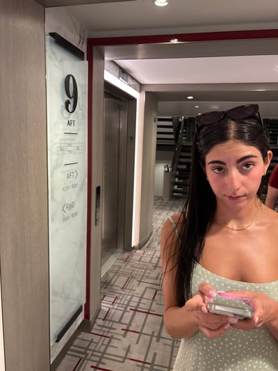 The scene depicts an interior hallway on Deck 9 of a cruise ship, consistent with a vessel potentially docked or operating near San Miguel de Cozumel, Mexico.
In the foreground, a young adult female with long, dark, wet hair pulled back and dark tortoiseshell sunglasses resting on her head, looks directly at the camera. She wears a light green, possibly floral-patterned, spaghetti-strap dress and a thin gold-colored chain necklace. Her face appears flushed. She holds a clear-cased smartphone horizontally in both hands, positioned as if typing or interacting with the screen. Her fingernails are painted pink. Partially visible on the far right, behind her, is the arm and shoulder of another individual wearing a dark red shirt.
To the left, a tall, vertical sign on a light wood-paneled wall displays "9 AFT", a simplified ship diagram, and cabin number ranges ("AFT > 9239-9329" and "FWD > 9100-9238"). The sign features a white marble-patterned background with dark numbering and text. Beyond this sign, two metallic elevator doors are visible in a recessed alcove, marked with a small "9". The entrance to this area is framed by a thin red trim. Further down the hallway, a dark stairwell is partially visible on the right side. The floor is covered with a geometric patterned carpet in shades of grey, white, and red. The ceiling is white with recessed lighting.
