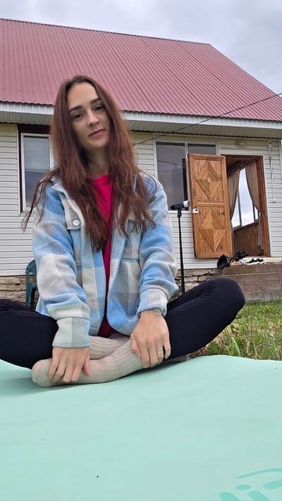 A young woman with long brown hair is sitting cross-legged on a light green mat outdoors. She is wearing a bright pink t-shirt and a blue and white plaid jacket. Her legs are crossed in front of her, and she is wearing light brown socks and black pants. Her hands are resting on her knees. Behind her is a light-colored house with a red metal roof. The house has a wooden double door that is open, revealing a dark interior. A black tripod with a camera on top is positioned to the left of the doorway. The ground is covered in green grass, and there are some stones and a plastic chair visible to the left of the woman. The sky is overcast.