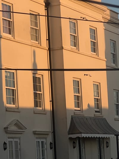 The image displays a multi-story, cream-colored building facade in Worthing, United Kingdom, illuminated by direct, low-angle sunlight.

The facade features multiple white-framed sash windows with visible glazing bars across at least three floors. A dark vertical downpipe is affixed to a structural column on the left side of the frame. On the middle floor, a small dark fixture, possibly a sensor, is mounted on the wall above two central windows. On the lower right, a dark, fluted metal awning with a scalloped edge extends over an entrance. Two black exterior wall lamps are mounted on the wall below this awning. To the left on the ground floor, a window or door unit is topped by a triangular pediment, and an adjacent unit has a flat architrave.

Prominent shadows are cast by the low sun. An elongated, human-like shadow is projected vertically across the upper left section of the building, spanning two floors. A second, shorter, broader shadow, consistent with a human head and shoulders, is cast on the lower right facade, above the awning. Additional shadows define architectural features such as ledges, window frames, and the downpipe.

Several dark, horizontal utility wires traverse the frame at various heights, partially obscuring the building. No individuals are physically visible within the image; their presence is solely indicated by the cast shadows.