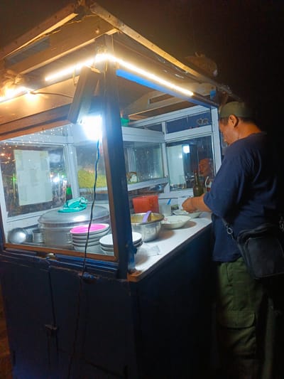 The image depicts a man preparing food at a food stall in Luwoo, Indonesia. The stall is illuminated by overhead fluorescent lights, casting a warm glow on the scene. The man, wearing a green cap and a dark blue t-shirt, is seen working with bowls and utensils, suggesting he is in the process of serving a customer.
The stall itself is made of wood and glass, with various food items and cooking equipment visible. A large metal pot sits on a shelf, likely containing a stew or soup. A stack of plates and bowls are neatly arranged, ready for use. In the background, there are glimpses of other food items, including what appears to be noodles in a container. The overall impression is one of a bustling street food vendor, providing a meal to the local community. The time of day appears to be evening or night, given the artificial lighting and the dark background.