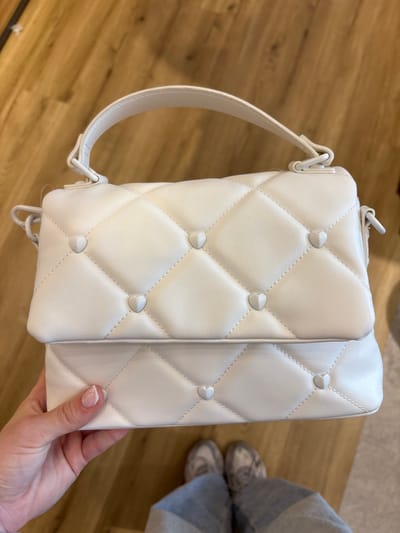 This is a top-down, close-up shot of a white quilted handbag. The handbag has a diamond-pattern quilting and is adorned with small, heart-shaped studs at the intersections of the diamond shapes. A white strap is attached to the bag. A person's hand with manicured nails is holding the side of the bag. In the background, a wooden floor is visible, and partially to the right, a person's legs in grey pants and silver shoes can be seen. The lighting suggests an indoor setting, possibly a retail environment or a home. The focus is on the handbag, highlighting its texture and decorative elements. There is no visible text. The scene appears casual and observational.