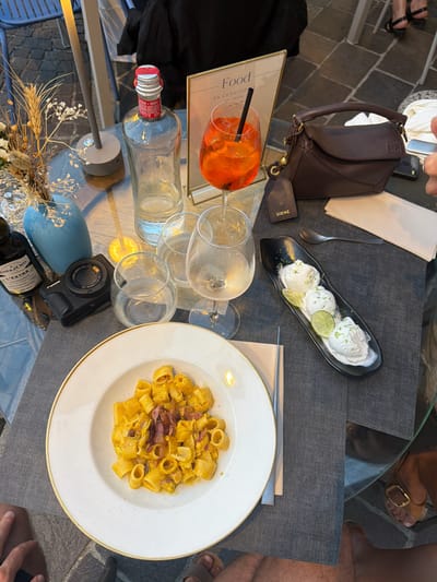 The overhead shot depicts a dining scene on an outdoor patio in Loveno, Italy, likely at dusk or during evening hours, with ambient lighting. In the foreground, a white ceramic plate with a golden rim holds a dish of pasta with a creamy yellow sauce and pieces of bacon. To the right, a black oblong dish contains two scoops of white ice cream garnished with lime slices and zest. A silver spoon rests beside the ice cream.
The table is covered with a dark gray textured placemat. Several wine glasses, some containing white wine, are scattered around the table. A bottle of clear liquid, possibly spirits, stands upright. Adjacent to the bottle is a table lamp with a concrete base. A small bouquet of dried flowers and wheat in a light blue vase sits to the left of the lamp. A black compact camera is placed on the table near the flowers.
A dark brown leather handbag, featuring the Loewe logo on a detached tag, is positioned to the right of the bottle and in front of a small stand displaying the word "Food." An orange cocktail with ice and a black straw sits in a stemmed glass. A folded white napkin and a silver utensil are also visible.
The lower half of the image shows the legs and feet of at least two individuals, suggesting they are seated at the table. One person's legs are visible in the bottom left, wearing dark shorts. Another person's legs and feet, wearing sandals with gold buckles, are visible in the bottom right. The background is blurred, hinting at a European-style streetscape with cobblestones and building facades.