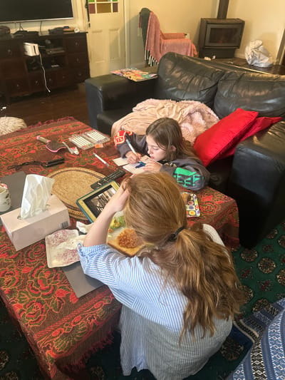 In a dimly lit living room, likely in Thora, Australia, two young girls are engaged in activities at a coffee table. The room is furnished with a large, black leather couch and a dark wooden entertainment unit. The coffee table is covered with a patterned fabric.

One girl, with long auburn hair, is seated and appears to be eating. In front of her is a plate with toast topped with baked beans and some avocado slices. She wears a blue and white striped shirt. She is resting her head on her hand.

The other girl, with brown hair, is also seated at the table, facing away from the camera. She is drawing or writing on a piece of paper with markers and a pen. She is wearing a grey hoodie with green lettering. A pink blanket is draped over the back of the couch behind her.

On the table, there are various items including a tissue box, a hairbrush, a remote control, a tablet device, a notebook, and a set of markers. The flooring is covered by a dark rug with a patterned design. The overall impression is of a casual, domestic setting where the children are occupied with creative pursuits and a meal.