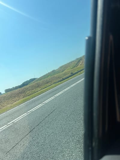 The image shows a slightly tilted view of a paved road with white lane markings, viewed from a vehicle. To the left of the road, a grassy embankment slopes upwards, with dry vegetation and scattered green patches. Further up the slope, a line of trees is visible, and beyond that, a clear blue sky with a sun flare in the upper left corner. On the right side of the image, a dark, vertical, and out-of-focus object partially obstructs the view, appearing to be part of the vehicle's interior or frame. The scene depicts a daytime outdoor environment, likely in a rural or semi-rural setting, given the natural landscape alongside the road. The specific location in the city of Marsum, Netherlands, cannot be determined from the visual information provided in the image.