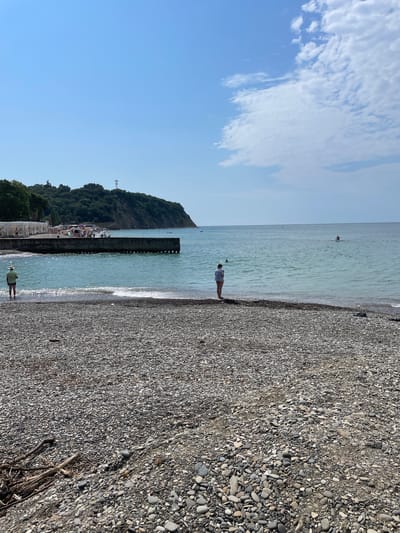 The image depicts a coastal scene in Krinitsa, Russia, likely during daylight hours under a clear blue sky with scattered clouds. The foreground is dominated by a pebble beach, with various sizes and colors of stones. A few pieces of driftwood are scattered on the pebbles.
In the mid-ground, the shallow turquoise water meets the shore with gentle waves. Two individuals are visible near the water's edge. One, wearing a straw hat and a light-colored shirt and shorts, stands facing the sea. Another, wearing a white top and shorts, stands looking out at the water.
Further out in the sea, several people are present. Some are swimming, while at least one individual is on a board, possibly a paddleboard or surfboard.
The background features a concrete pier extending into the sea. Beyond the pier, a densely forested hill rises, with a cellular tower visible near its summit. Along the shoreline in the distance, there are some structures that appear to be beach umbrellas or cabanas, and a number of people are gathered. The overall atmosphere is one of a calm day at the beach.