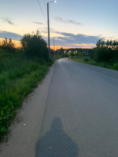 A paved road curves through a rural landscape at dusk. On the left, a grassy verge borders the road, with a line of trees and bushes in the distance. On the right, the road stretches ahead, lined with streetlights that are beginning to illuminate. The sky is a gradient of pale blue and soft orange, with scattered clouds. A shadow of a person, presumably the photographer, is cast on the road in the foreground. The overall scene suggests a quiet, possibly isolated location, typical of a village or countryside setting. The mention of Ganino, Russia, places the scene in a specific geographical context.