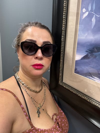 A woman with graying hair pulled back, wearing large dark sunglasses, bright pink lipstick, and a floral print top, poses for a selfie. She is adorned with multiple layered necklaces, including one with a cross pendant. She is positioned next to a framed painting with a dark, ornate frame. The background is a plain gray wall. The scene appears to be indoors, possibly in a home or gallery.