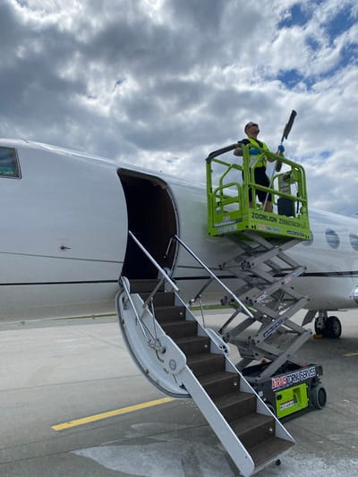This outdoor photograph, taken in Skulte, Latvia, captures a worker on a scissor lift next to a white private jet on an airport tarmac under a cloudy sky. The image is oriented vertically, showing a low-angle perspective looking up at the aircraft and the worker.

The central subject is a male worker, wearing a high-visibility vest, dark clothing, sunglasses, and gloves, positioned within the platform of a lime green scissor lift. He is holding a long-handled tool, possibly for cleaning or maintenance, and appears to be working on the aircraft's fuselage. The scissor lift, a ZOOMLION ZS0607ACW-Li model, has "ATRAIS TORNU SERVISS" and contact information ("www.ATS.LV", "27 555 777") printed on its side.

The large white aircraft, likely a private jet, dominates the right side of the frame, with its main passenger door open, revealing a view into the cabin. Adjacent to the aircraft's open door are mobile airstairs, allowing access to the interior. Partially visible near the airstairs is another piece of ground support equipment, also branded with "ATRAIS TORNU SERVISS" and its contact details, suggesting it belongs to the same service company.

The setting is an airport apron, characterized by a grey concrete surface with yellow demarcation lines. The time of day is daylight, with diffused lighting from the overcast sky. The overall scene depicts routine maintenance or servicing of an aircraft. Visible text on the aircraft door includes "OFF", "NO HAND GRIP", and "CAUTION".