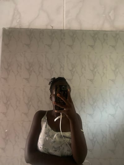 The image shows a young woman taking a mirror selfie in what appears to be a bathroom or tiled room. She is holding a smartphone to capture the reflection of herself. The background consists of tiled walls with a marble-like pattern. She is wearing a light-colored, floral-patterned camisole or tank top. Her hair is dark and styled in curls. The lighting suggests it could be daytime, possibly indoors with artificial light. The reflection of the phone shows text which is reversed due to the mirror, and it's difficult to discern clearly but appears to have some writing. The overall mood is casual and personal, as is typical of a selfie. There are no obvious indicators of weather or specific time of day beyond general indoor lighting. The location context provided is Wauro Leggal, Nigeria.