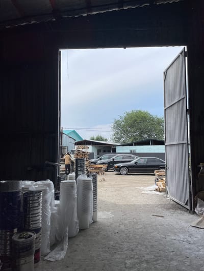 The image captures a view from inside a dimly lit industrial building, looking out through a large open doorway. The foreground is dominated by stacks of various containers, some metallic and cylindrical, others wrapped in white plastic. A person in a light-colored shirt and dark pants is visible just outside the building, standing near a truck and appearing to be interacting with it.
Beyond the immediate area, the scene opens up to an outdoor yard. Two black sedans are parked side-by-side. Pallets of lumber are stacked in the yard. In the background, several buildings with differing architectural styles and colors are present, including one with a distinctive teal-colored roof. A large tree with dense foliage stands behind some of the buildings. The sky above is overcast with a light grey hue. The overall impression is an industrial or storage area, likely in a rural or semi-urban setting within the village of Kenen Azirbayev, Kazakhstan.