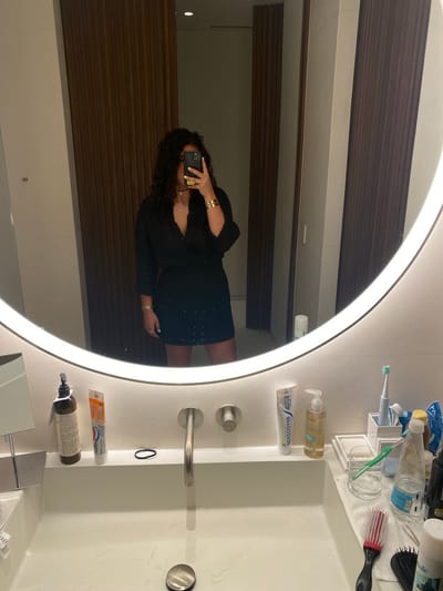 A woman is taking a selfie in a bathroom mirror. The mirror is round with a lit border. The woman is wearing a black dress and jewelry. The bathroom countertop is white and cluttered with various toiletries, including toothpaste, toothbrushes, soap, and other personal care items. The wall behind the mirror is covered with horizontal wooden panels. The photo does not provide any information to suggest that this photo was taken in El Caracol, Mexico.