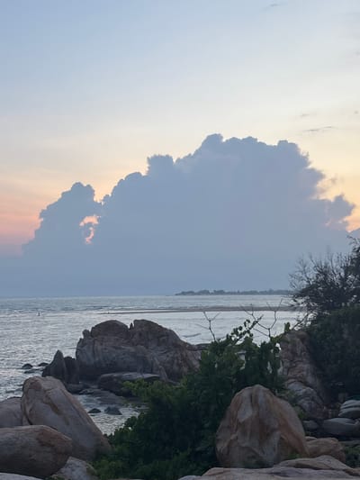 This photograph captures a serene coastal landscape at dusk, likely in Thuận Thành, Vietnam. The sky is painted with soft hues of peach and pale blue, transitioning into a light gray near the horizon. Dominating the center of the frame is a large, dramatic cloud formation, illuminated from behind by the setting sun, with a portion of the light peeking through a gap in the clouds.
The foreground is characterized by a rugged, rocky shoreline. Large, weathered boulders of varying sizes are scattered across the scene, some partially submerged in the calm, gray water of the ocean. Lush green foliage and sparse, dry branches are interspersed among the rocks, adding texture and depth. The ocean itself appears tranquil, with gentle ripples reflecting the muted light of the sky. In the distance, a thin strip of land is visible, possibly a sandbar or the distant coastline, with a few scattered structures or trees hinting at human presence. A solitary figure can be observed standing in the shallow water on the left side of the frame, appearing as a small, dark silhouette against the bright water. The overall impression is one of natural beauty and quiet contemplation, characteristic of a peaceful evening by the sea.