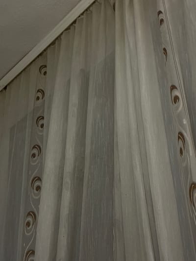 The image displays a close-up, upward-angled view of light-colored, translucent curtains in an indoor setting in Vlahovo, Bulgaria. The fabric exhibits fine vertical striations and is gathered into multiple soft, vertical folds. A decorative vertical band, present on at least two sections of the curtain, features a repeated pattern of stylized brown and white spiral or swirl motifs interconnected by a thin, wavy white line. Above the curtains, a white curtain rod or cornice is visible, connected to a white, textured ceiling in the upper left quadrant of the frame. The lighting is even and diffused, creating soft shadows within the curtain folds. No individuals or specific actions are depicted.