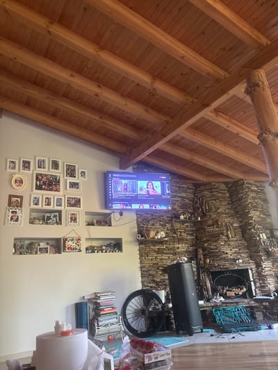 The image shows an interior scene of a room with a high, vaulted wooden ceiling. The walls are adorned with a collection of framed photographs, creating a gallery effect. Some of the frames are white, while others appear to be made of wood or metal. A flat-screen television is mounted on the wall and is displaying content, possibly from YouTube, with visible Russian text on the screen.
Adjacent to the television is a section of the wall covered in stacked, irregular stones, giving it a rustic texture. Above this stone-covered section, various objects are visible, including a set of tools in a green case, a black storage case, and a bicycle wheel. There are also shelves with decorative items and books.
The overall impression is one of a lived-in and personalized space. The collection of photos suggests a focus on family and memories. The combination of natural wood, stone, and collected items creates a cozy and eclectic atmosphere. The lighting appears to be artificial, indicating it is likely indoors and not dependent on natural daylight. The scene does not provide strong cues about the time of day or weather. The location context provided is Prolesha, Bulgaria, which could imply a rural or suburban setting.