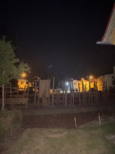 This image, taken at night in Jablonné nad Orlicí, Czechia, shows a dimly lit outdoor scene. The foreground is dominated by dark green foliage on the left and a grassy area on the right, with a rough wooden fence structure running through the middle. The fence appears to be constructed from uneven planks and posts, possibly part of a playground or an unfinished construction.

Beyond the fence, in the background, are several buildings illuminated by artificial light, suggesting a residential or urban area. One building has a prominent light on the second floor, and other windows are lit. A streetlamp is visible, casting a warm glow. Scattered on the ground near the fence are several stakes, some with colored tips (red and yellow), possibly marking out an area or indicating construction progress.

The overall impression is of a quiet, somewhat rural or suburban setting at night. The lighting is ambient, with the primary illumination coming from the buildings and the streetlamp. The dark sky and limited light sources create a somewhat subdued atmosphere. There is no visible text in the image. The crops provided highlight details of the building facades, the construction of the fence, and the stakes in the ground.