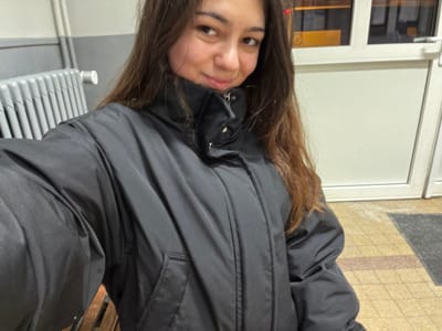 A young woman with long brown hair poses indoors. She wears a black jacket. Behind her is a white door and a radiator. The image is rotated, with the woman appearing upside down. Tiled flooring is also visible.