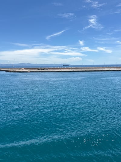 The image displays a serene and expansive coastal view on a clear, sunny day, likely from an elevated perspective. The scene is dominated by three main horizontal bands: sky, a distant landmass with a pier, and the sea.

In the upper portion of the frame, a vibrant blue sky stretches across, adorned with scattered, wispy white clouds and some larger, fluffier cumulus clouds, indicating fair weather. Below the sky and central to the composition, a very long, straight pier or breakwater extends horizontally from left to right. This substantial structure, likely made of concrete or stone, is lined with numerous evenly spaced poles, possibly streetlights or navigational aids. Behind this pier, in the distant background, a hazy, mountainous coastline or rolling hills rise, appearing in muted blue tones under the bright sky. A dark-hulled cargo ship or tanker is visible in the waters behind the pier, suggesting active maritime operations typical of a port.

The lower part of the image is filled by the calm, deep blue water of the sea. Its surface is textured with countless small ripples and gentle waves, reflecting the sunlight in sparkling glints, particularly noticeable towards the right side of the frame. The water appears tranquil and clear.

The overall impression is one of peaceful grandeur combined with the industriousness of a port. Given the location context of Piombino, Italy, the scene likely depicts its significant harbor area. The time of day is clearly daytime, with bright sunlight. No people are directly visible, and there is no discernible text within the image.