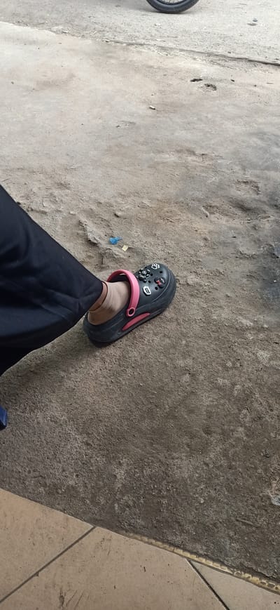 A person's left foot, clad in a black Croc-style sandal with pink accents and various charms, rests on a concrete surface. The sandal is decorated with several small charms, including a paw print, a cartoon character, and what appear to be letters or symbols. The person is wearing a light-colored ankle sock and black pants. The setting appears to be outdoors, with a rough, textured concrete ground. In the background, a portion of a motorcycle wheel is visible. The lighting suggests it is daytime, possibly overcast given the diffused light. The scene is simple and casual, with no strong emotional cues apparent. No discernible text is visible beyond the charms on the shoe.