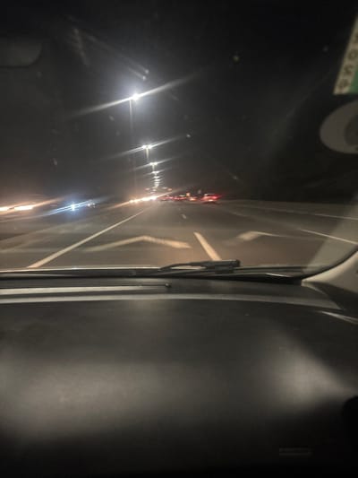 A nighttime view from a car window shows a highway with bright headlights and taillights stretching into the distance. Lanes are marked with white lines. The car's wiper blade is visible. The side panel of the car is featured on the right. A reflection is visible from the car window in the dark area to the left.