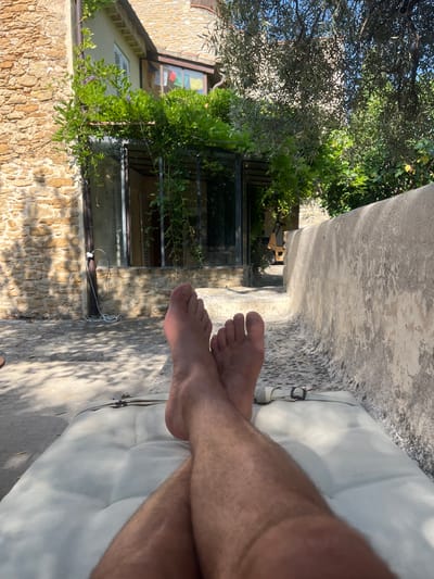 The image depicts a personal perspective of someone relaxing outdoors in Le Castellet, France. The foreground is dominated by the lower legs and feet of an individual, who is lying on a light-colored padded surface. Their legs are crossed at the ankles, and their feet are bare.

The background reveals a rustic setting with a stone building constructed from rough-hewn, light brown stones. A large glass-paned door or window is visible on the building, partially obscured by abundant green foliage and climbing vines with small purple flowers. Above the windows, a section of the building's roof with terracotta tiles is visible, along with a blue sky.

To the right of the building, a weathered, textured concrete wall curves along the scene, creating a natural seating or lounging area. The ground in front of the building and extending towards the concrete wall is paved with irregular stone or concrete slabs, dappled with sunlight and shadow. Mature trees with silvery-green leaves, likely olive trees, cast shadows over parts of the scene, contributing to the warm, sunlit atmosphere. The overall impression is one of peaceful, leisurely repose in a Mediterranean environment.