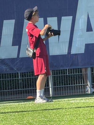 A person wearing a red shirt and shorts is standing by a fence, taking a photo with a camera in Fentange, Luxembourg. They are wearing a dark-colored hat, socks, and athletic shoes. A small bag is hanging from their shoulder. The backdrop includes a large blue sign with white geometric shapes and a green grassy area. The scene is outdoors and appears to be during daylight hours.