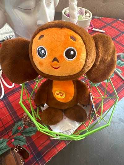 An image captured in Matveevskoe, Russia, displays a plush Cheburashka toy positioned centrally within a lime green, geometric wire-frame basket. The toy features a brown body, large round brown ears, an orange face with large black eyes and white reflections, an upturned red mouth, and a small V-shaped brown nose. An orange patch with a yellow musical note icon is visible on its chest. A small tag hangs from the toy's right side. The basket rests on a red and black plaid table runner, which is decorated with green holly leaves, red berries, and brown pine cones. The surface beneath the runner is dark. In the background, to the left, a portion of a large white ceramic decorative object, possibly a stylized face, is visible. To the upper right, a small white pot containing colorful stones and a white textured decorative element is present. A brown leather wallet or pouch is partially visible in the bottom left corner, resting on the plaid runner. No individuals are depicted, and all objects are stationary.