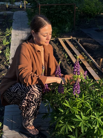 A woman is crouching down next to a cluster of purple lupine flowers in a garden. She is wearing a brown oversized shirt and patterned pants. The garden has a stone pathway and raised garden beds with young plants. The scene is lit by the warm light of the sun, casting shadows across the pathway. The location is identified as Kansk, Russia.