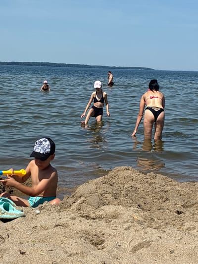The image depicts a sunny day at a beach in Rewa, Poland. In the foreground, a young boy wearing a black and white patterned cap and blue swim trunks is playing in the sand with a yellow toy shovel and a blue bucket. To his left, a woman in a black bikini is partially submerged in the water, her back to the camera, with her hand touching the water's surface. Further in the water, another woman wearing a black bikini and a white cap is visible. In the distance, a person is swimming in the water, and the shoreline stretches out with trees in the background. The water appears calm, with gentle ripples, and the sky is a clear, bright blue, suggesting pleasant weather and a midday time frame. The scene evokes a sense of summer leisure and enjoyment at the seaside. There is no visible text in the image.