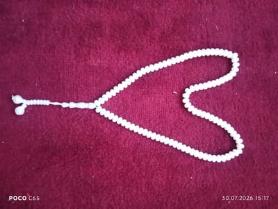 A string of small, white, round beads is laid out in a heart shape on a dark red, textured surface, likely fabric. The beads appear to be made of a smooth, possibly plastic or polished stone material. The left side of the string has a tassel-like arrangement of white beads. The image also has text overlays: "POCO C65" in the bottom left corner and "30.07.2026 15:17" in the bottom right corner. The background is a uniform, deep red with a slight fuzzy texture, suggesting a blanket or carpet. There are no people visible in the image. The scene does not contain any discernible elements that would indicate a specific location within the city of Beruniy, Uzbekistan.