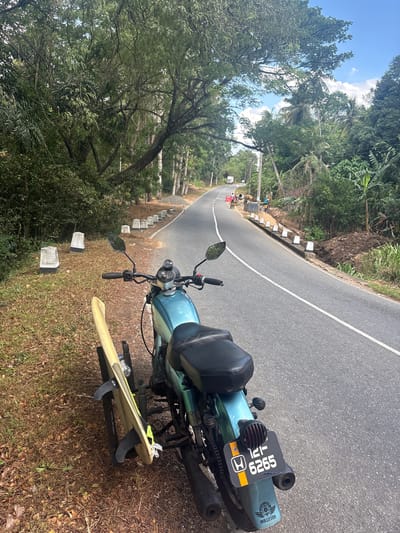 The image shows a turquoise motorcycle parked on the side of a paved road in a rural, tropical setting. The motorcycle is facing away from the camera and has a surfboard strapped to its side. The surfboard is pale yellow with black fins. A license plate is visible on the back of the motorcycle, displaying "121-6265". The road has a white line down the center and curves to the left. To the left of the road, there is dense green vegetation and trees, with a clear blue sky visible above. To the right of the road, there is a grassy embankment.
In the distance along the road, there appear to be some people and construction equipment, possibly indicating road work or a landslide. There are also several concrete markers along the side of the road. The scene suggests a sunny day, likely late morning or early afternoon given the brightness and the shadows. The lush vegetation points towards a warm, humid climate. The presence of the surfboard and motorcycle suggests travel and a potential recreational activity. The overall mood is one of adventure and exploration in a scenic, natural environment.