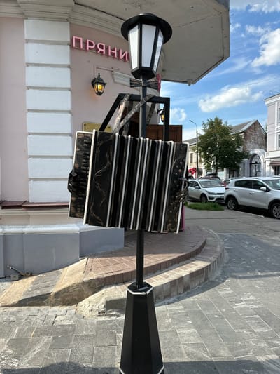 An outdoor scene in the city of Putyatino (Semivragovskaya S/A), Russia features a street with buildings and parked cars. In the foreground, a large, decorative accordion sculpture is mounted on a lamppost. The accordion is black with white keys and gold decorative elements. Behind the sculpture, a building with pink siding and white columns is visible, with a sign that reads "ПРЯНИ" in pink lettering. A vintage-style lantern hangs from the building. The street is paved with stone tiles, and several white cars are parked on the right side of the image. Trees and other buildings are visible in the background under a partly cloudy sky.