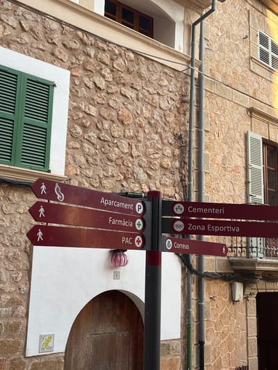 This image depicts a street scene in Fornalutx, Spain, characterized by traditional stone architecture and a prominent directional signpost.
In the center of the frame, a dark grey and maroon multi-armed signpost stands, indicating six local destinations. Three maroon arrow-shaped arms point to the left: "Aparcament" (Parking) with a 'P' symbol and a pedestrian icon, "Farmàcia" (Pharmacy) with a green cross and a pedestrian icon, and "PAC" (Primary Care Center) with a red cross and a pedestrian icon. Three corresponding maroon arrows point to the right: "Cementeri" (Cemetery) with three cross symbols, "Zona Esportiva" (Sports Zone) with the Olympic rings symbol, and "Correus" (Post Office) with a stylized envelope symbol and a pedestrian icon.
Behind the signpost, two buildings with rough-hewn stone facades are visible. The building on the left features a rectangular window with closed, dark green louvered shutters in the upper-left quadrant. Below the sign, a white-painted archway leads to a wooden door with visible studs. A small, square ceramic tile depicting a yellow bird on a branch, labeled "CAN MOSSO," is affixed to the white wall next to the door. Above the archway and below the lowest left-pointing sign, a pink, ribbed, opaque glass or ceramic lamp fixture is mounted, with the number "1" below it.
The building on the right, partially visible, also exhibits a stone facade. It features a window with partially open, light grey/white louvered shutters. A dark grey downspout runs vertically along the wall of this building, accompanied by multiple electrical conduits and wires. The scene is illuminated by daylight, suggesting an outdoor daytime setting, with no individuals present.