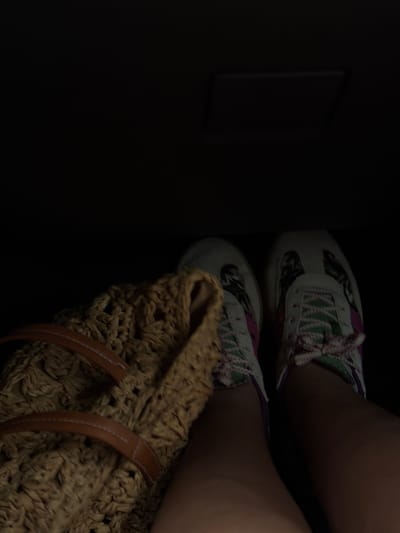 A person's legs are visible, wearing patterned sneakers and resting on a dark surface, possibly the floor of a vehicle. A woven straw bag with leather straps is placed to the left of the legs. The background is dark and out of focus, with a rectangular object dimly visible in the upper portion of the image. The scene appears to be indoors, possibly in Groisy, France, given the context provided.