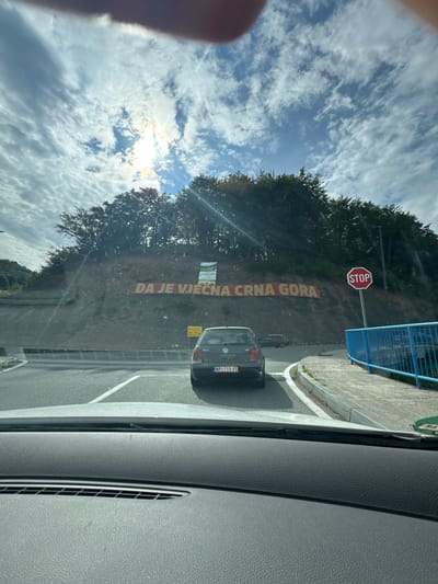 The image shows a view from inside a car looking out towards a hillside with a large red banner on it. The banner reads "DA JE VJECNA CRNA GORA", which translates to "Long live Montenegro". A stop sign is visible to the right of the banner. The landscape is hilly and covered in trees. A grey Volkswagen Golf is parked on the road in front of the banner. The sky is cloudy with the sun shining through. The location is identified as Lugovi, Montenegro.