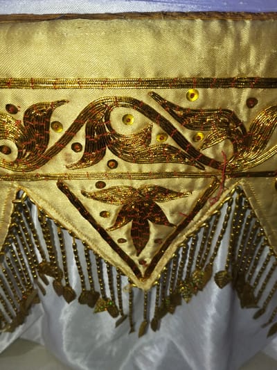 The image is a close-up of an ornate textile, likely a garment or decorative fabric, featuring intricate gold embroidery and beadwork. The primary fabric appears to be a pale gold or beige silk with a visible weave. A horizontal band of layered gold metallic trim runs across the upper portion of the image, above a decorative motif. This motif consists of swirling, curvilinear patterns embroidered in gold thread, interspersed with small, reddish-brown sequins and tiny gold sequins. Beneath this upper motif, a triangular section of the pale gold fabric is bordered by gold trim. From the lower edge of this triangle hang numerous strands of gold beads, each tipped with a small, leaf-shaped gold embellishment. The textile below this beaded fringe is a smooth, white or light gray fabric, likely satin, which drapes loosely. The lighting in the image is somewhat harsh, creating highlights and shadows that emphasize the textures and dimensionality of the embroidery. There are no people or environmental elements visible beyond the textile itself, and no specific location in the city of Cottrueng, Indonesia can be determined from the image.